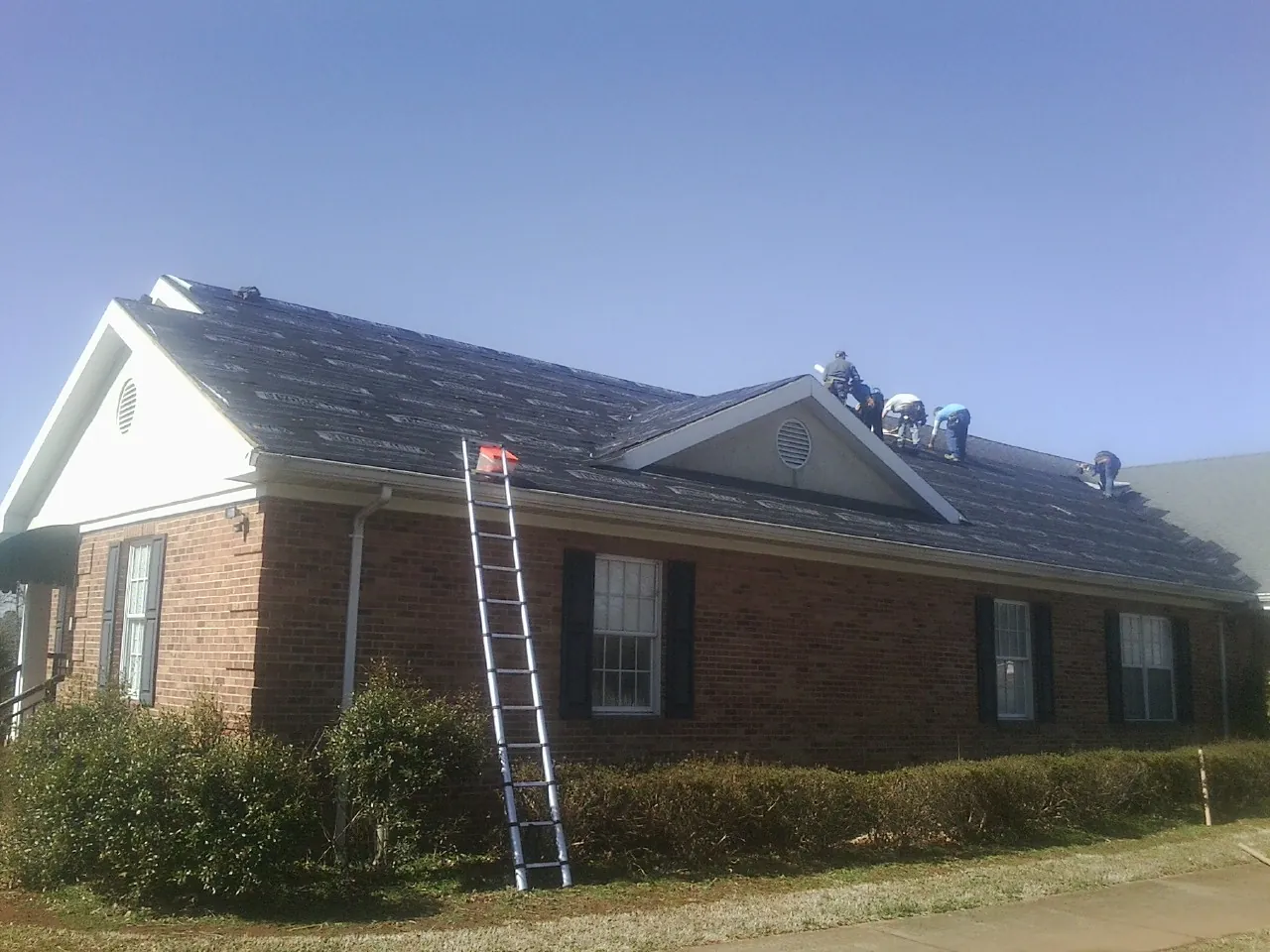 Crew tearing shingles off a brick ranch roof with a ladder set — Metal Roof Repair work by our crew in Riegelwood