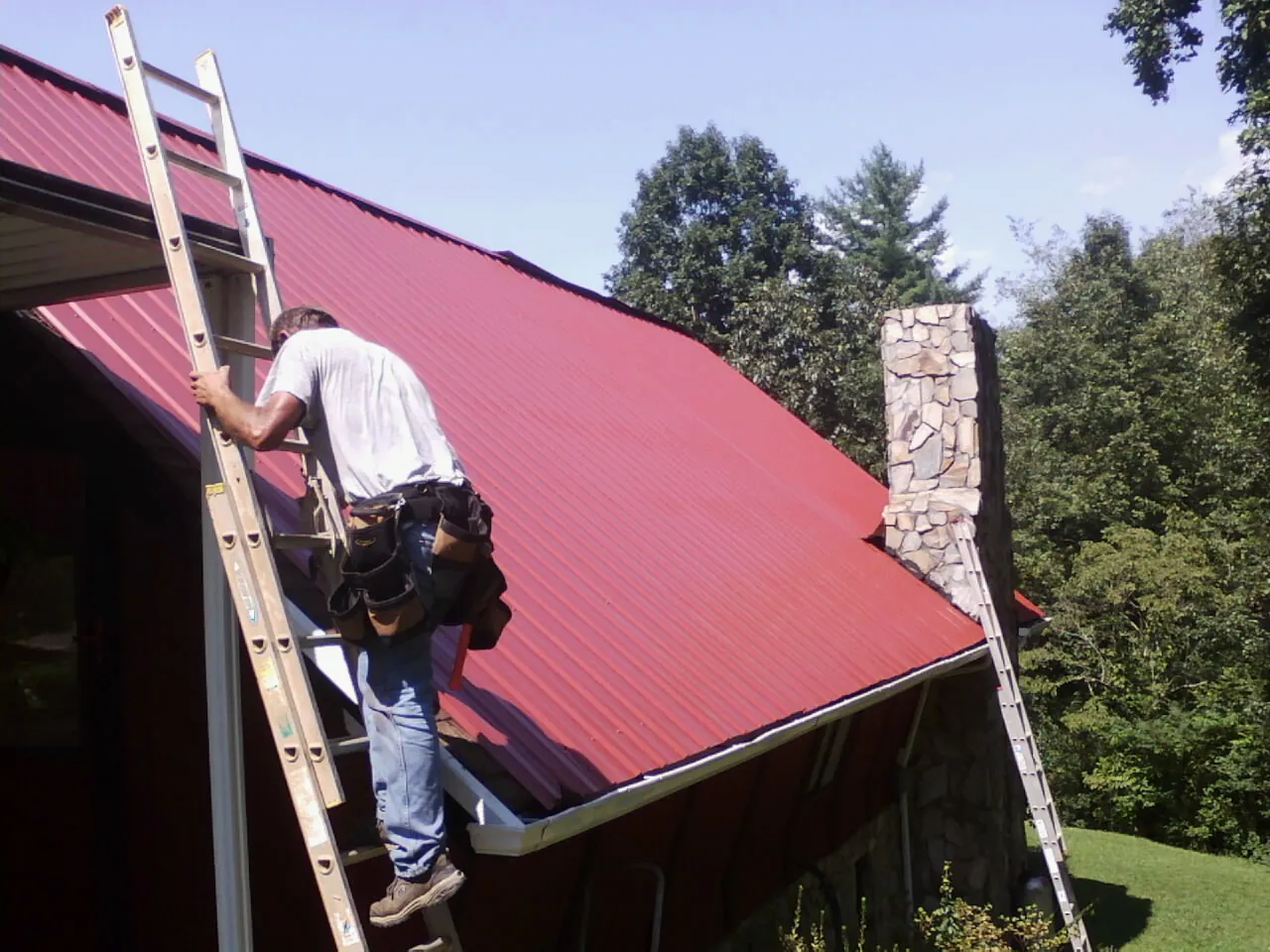 Roofer on a ladder beside a red metal roof and stone chimney — Soffit & Fascia Repair by Roofing Brothers in Riegelwood